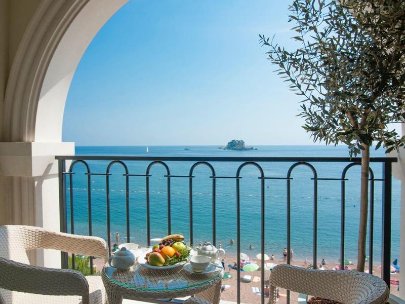 Balkon mit runder Bogenöffnung, zwei Stühle, Tisch mit Obst, Blick auf ruhiges Meer und klaren Himmel.