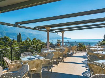 Terrasse mit Tischen und Stühlen bietet Blick auf Meer und Berge bei klarem Himmel.