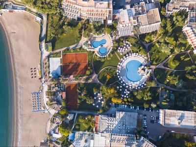 Luftaufnahme eines Hotels mit zwei großen Pools neben einem Sandstrand und umliegenden Gebäuden.