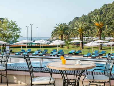 Poolbereich mit Sonnenschirmen, Liegestühlen, Tischen, Stühlen und Palmen im Hintergrund.