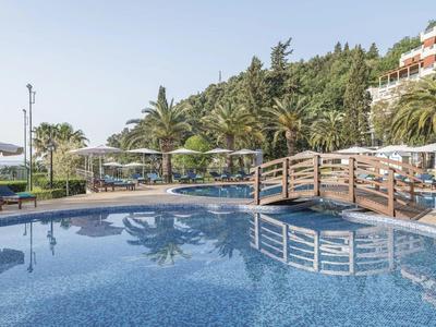 Großer, blauer Pool mit Brücke, Liegestühlen und Palmen unter klarem Himmel bei Hotelgebäude.