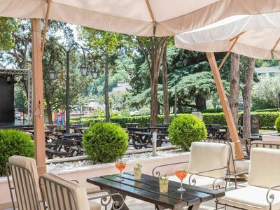 Outdoor-Café mit weißen Sonnenschirmen, Glasplatten-Tischen und gepolsterten Stühlen unter Bäumen.