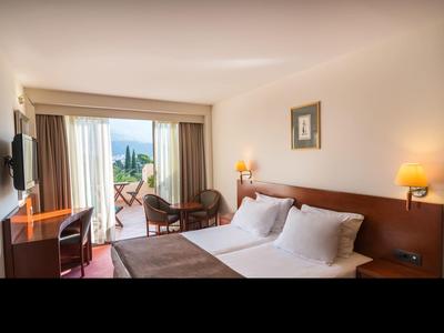 Hell eingerichtetes Hotelzimmer mit Doppelbett, Schreibtisch und Balkon mit Sitzgelegenheit