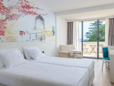 Helles Hotelzimmer mit zwei Einzelbetten, Balkon und Sitzbereich mit Stuhl und Sessel.