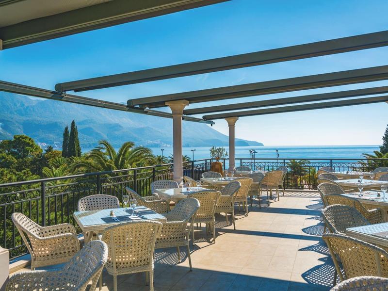 Terrasse mit Tischen und Stühlen bietet Blick auf Meer und Berge bei klarem Himmel.