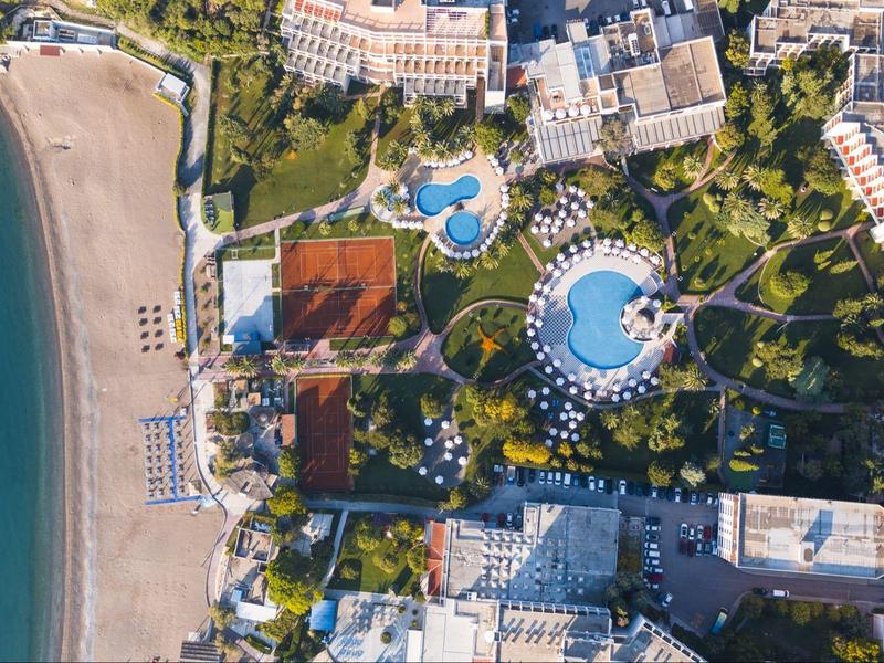Luftaufnahme eines Hotels mit zwei großen Pools neben einem Sandstrand und umliegenden Gebäuden.