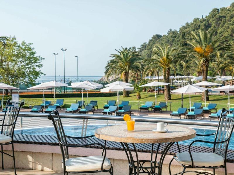 Poolbereich mit Sonnenschirmen, Liegestühlen, Tischen, Stühlen und Palmen im Hintergrund.