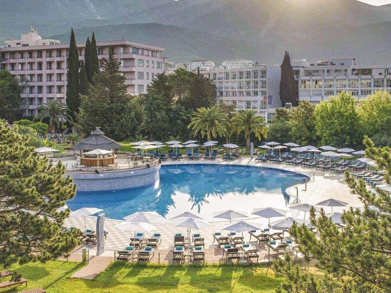 Großer Außenpool mit Sonnenliegen in einer Hotelanlage, umgeben von Bäumen und Bergen im Hintergrund