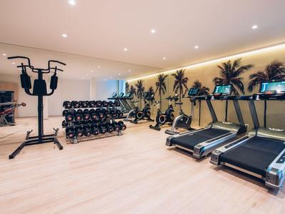 Moderner Fitnessraum mit Hanteln, Laufbändern und stationärem Fahrrad vor Palmenwand.