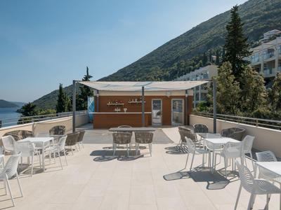 Taras na zewnątrz z białymi stołami i krzesłami, widokiem na morze i budynki na wzgórzu.