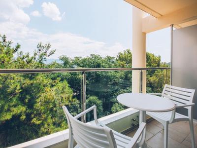 Balkon z białymi krzesłami i stołem z widokiem na zielony las pod częściowo zachmurzonym niebem.