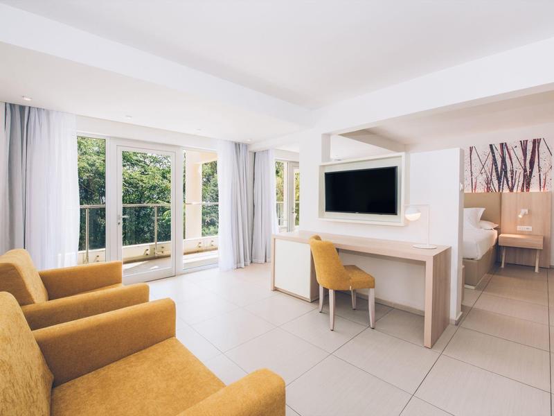 Jasny pokój hotelowy z dużymi oknami, żółtymi fotelami i biurkiem z telewizorem.