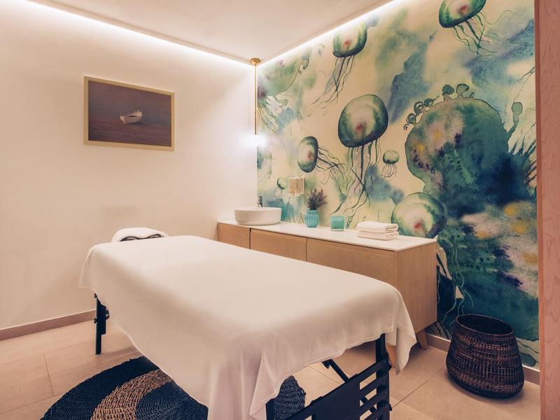 Przytulny pokój zabiegowy z łóżkiem do masażu i artystycznym wzorem na ścianie.
