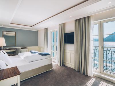 Bright hotel room with large bed, desk, TV, and balcony with mountain view.