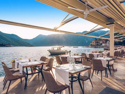 Restaurant terrace by the water with tables, chairs, and mountain view in sunny weather.