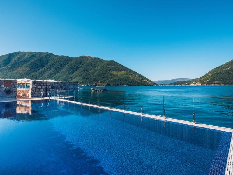 Infinity pool overlooking hilly landscape and clear blue water under clear sky.