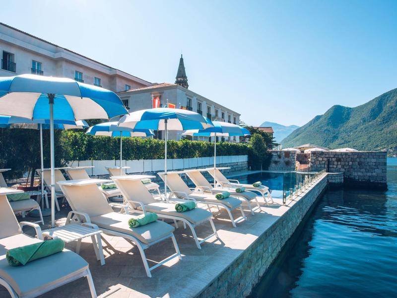 Sun loungers with blue umbrellas by a hotel pool with mountain views.