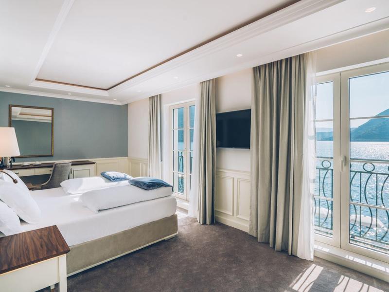 Bright hotel room with large bed, desk, TV, and balcony with mountain view.