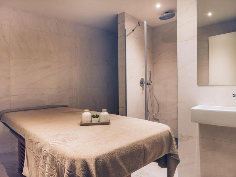 Treatment room with massage table, modern shower area, and sink in soft beige and cream tones.