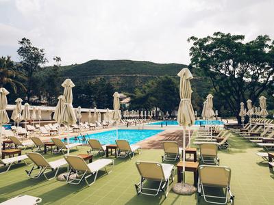 Großer Hotelpool mit vielen Liegestühlen und geschlossenen Sonnenschirmen vor bewaldetem Hügel.