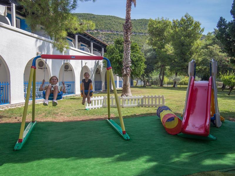 Spielplatz mit zwei Schaukeln und einer roten Rutsche in einem grünen Garten neben einem weißen Gebäude.