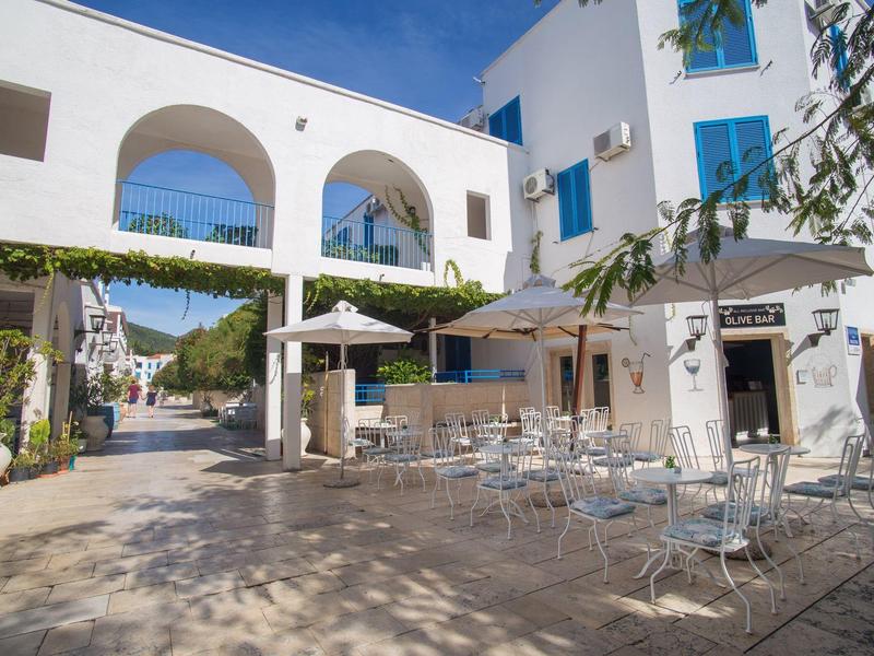 Innenhof eines weißen Hotels mit blauen Fenstern, Außensitzbereich und Arkaden unter blauem Himmel.