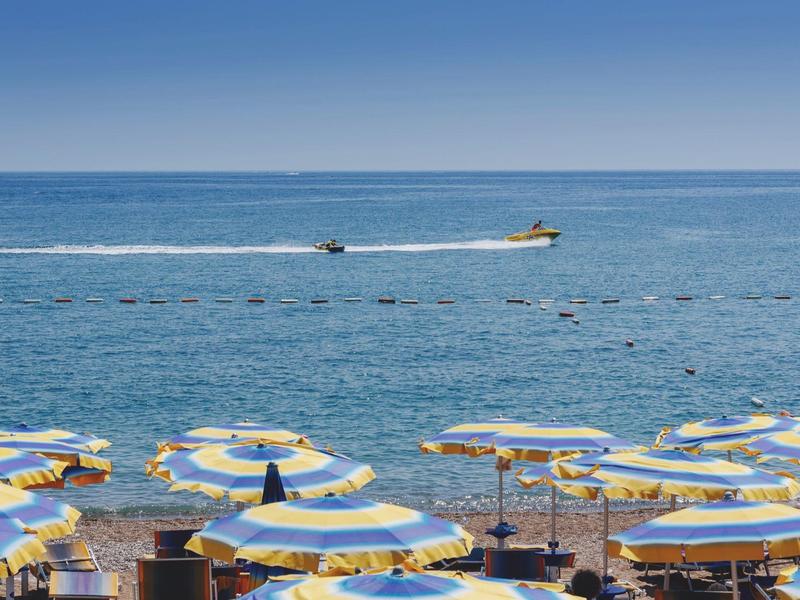 Gelb-blaue Sonnenschirme am Strand, dahinter blaues Meer mit zwei Schnellbooten auf dem Wasser.