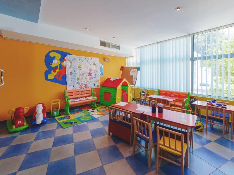 Kindergartenraum mit bunten Möbeln, Spielzeug, Teppichen und großen Fenstern für Tageslicht.