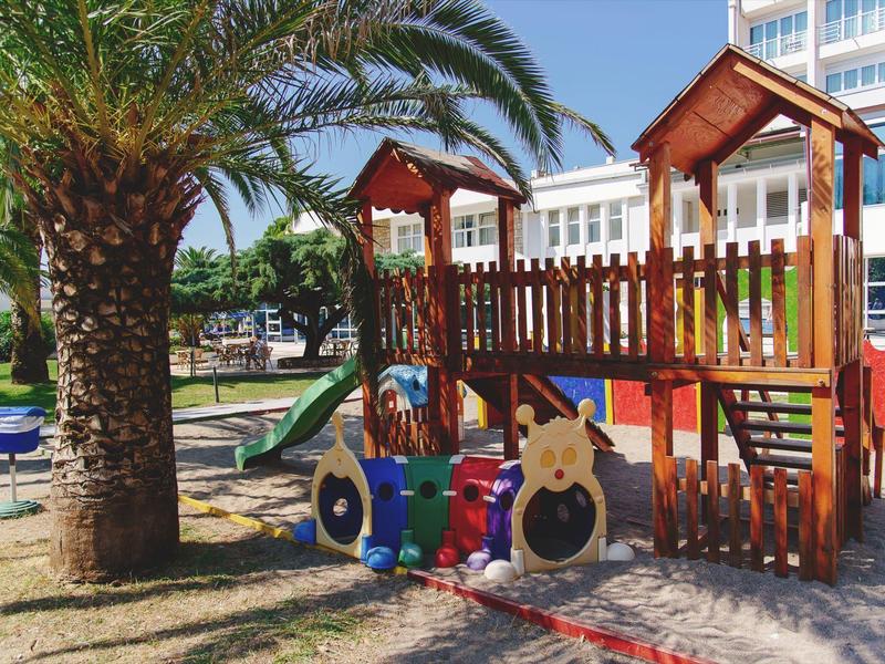 Bunter Spielplatz mit Rutsche, Klettergerüst und einer Palme bei sonnigem Wetter.