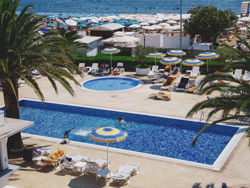 Schwimmbad mit blauer Wasserfläche, Sonnenschirmen, Palmen und Meerblick im Hintergrund.