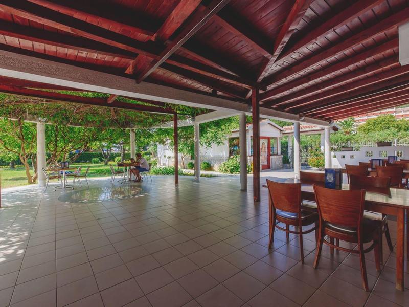 Überdachte, offene Terrasse mit Holzdach, Fliesenboden und mehreren Holztischen und -stühlen.