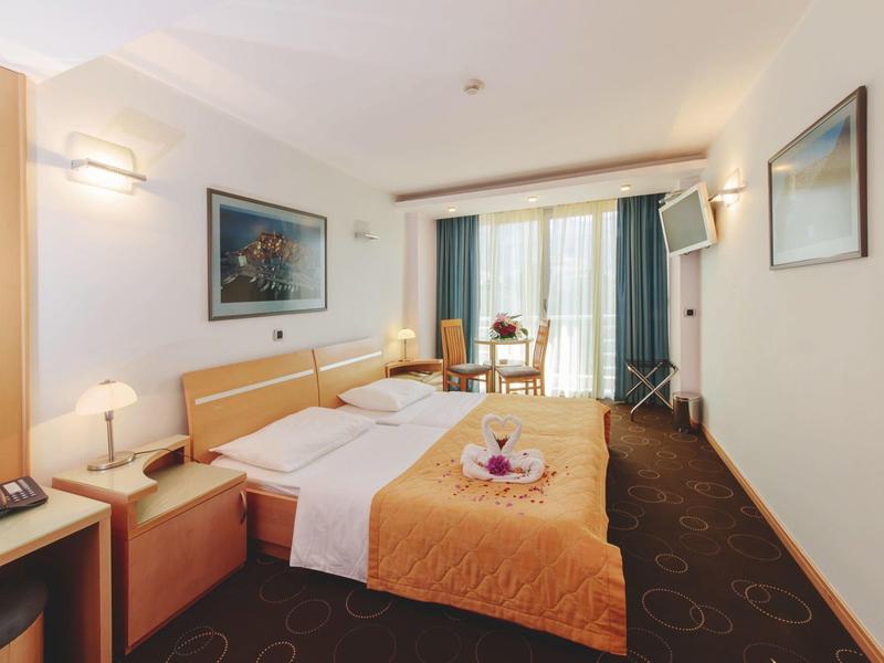 Helles Hotelzimmer mit großem Bett, orangefarbener Decke, Schreibtisch und Balkon mit Vorhängen.