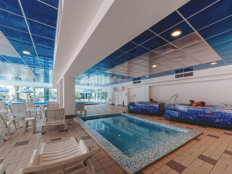 Innenpoolbereich mit gemustertem Becken, blauer Decke, weißen Liegen und Sitzbereichen.