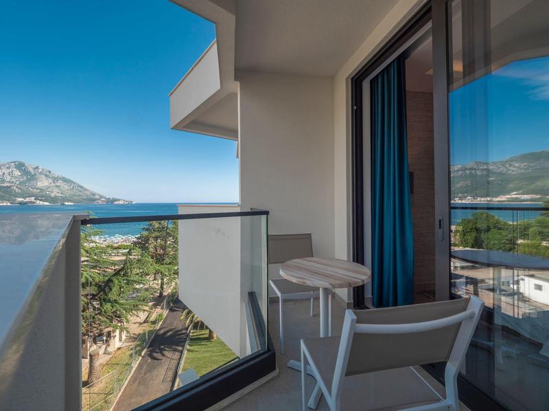 Balkon mit weißem Geländer, Stuhl und Tisch, Blick auf Meer, Bäume und Berge bei blauem Himmel.