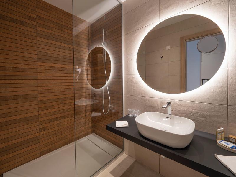 Moderne Dusche mit Holzfliesen, runder beleuchteter Spiegel und weißes Aufsatzwaschbecken auf schwarzer Ablage.