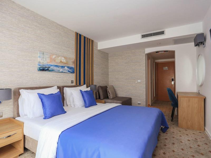 Helles Hotelzimmer mit Doppelbett, blauer Bettdecke, Nachttischen und Schreibtisch am Fenster.