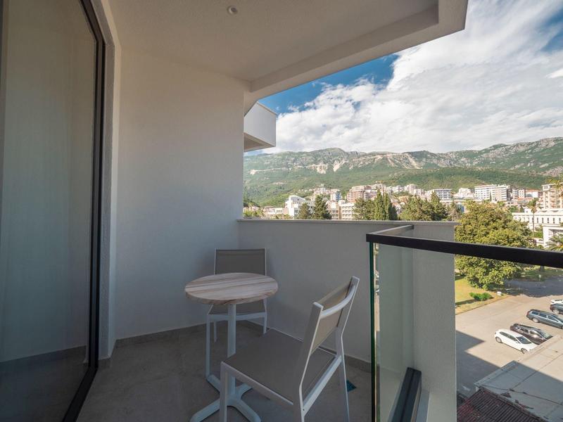 Kleiner Balkon mit rundem weißen Tisch, Stuhl, Glasgeländer und Blick auf Berge und Stadt.