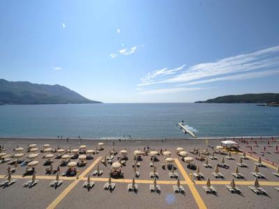 Leerer Strand mit Sonnenschirmen und einem Steg ins ruhige blaue Meer bei klarem Himmel.
