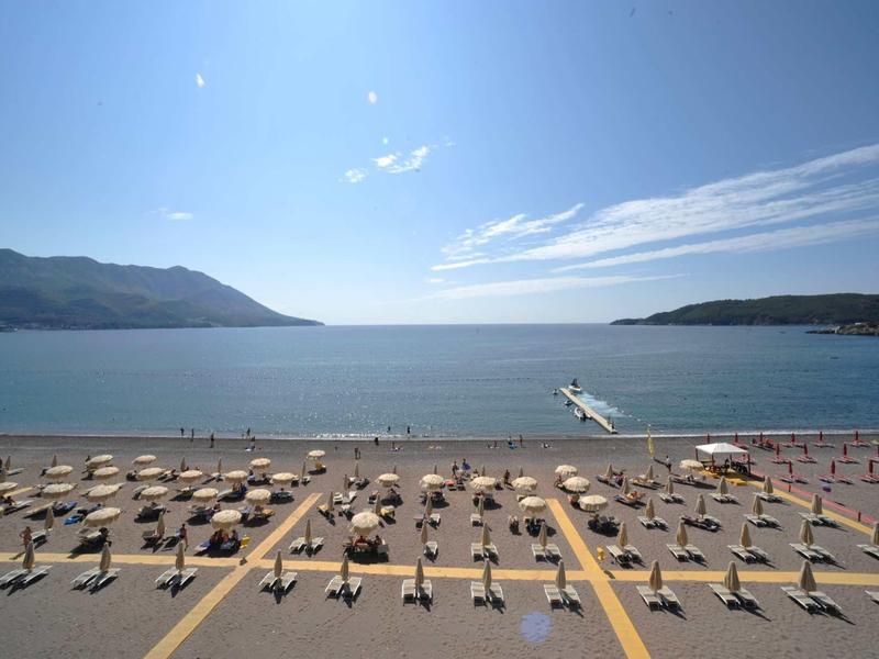 Strand mit vielen gelben Sonnenschirmen, Sand, blauer See und Himmel mit wenigen Wolken.