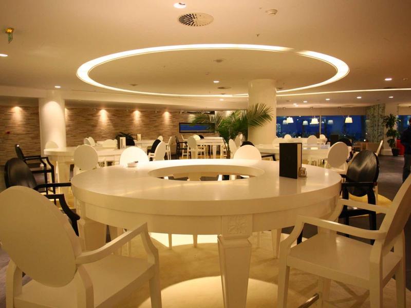 Moderne, helle Lounge mit runden weißen Tischen, beigen Stühlen und warmem Deckenlicht.