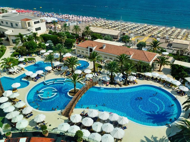 Großer Hotelpool mit runden Formen, Palmen und weißen Sonnenschirmen am Meerufer.