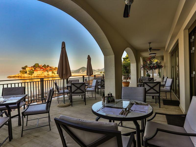 Überdachte Terrasse mit Tischen und Stühlen, Blick auf Wasser und Gebäude bei Sonnenuntergang.