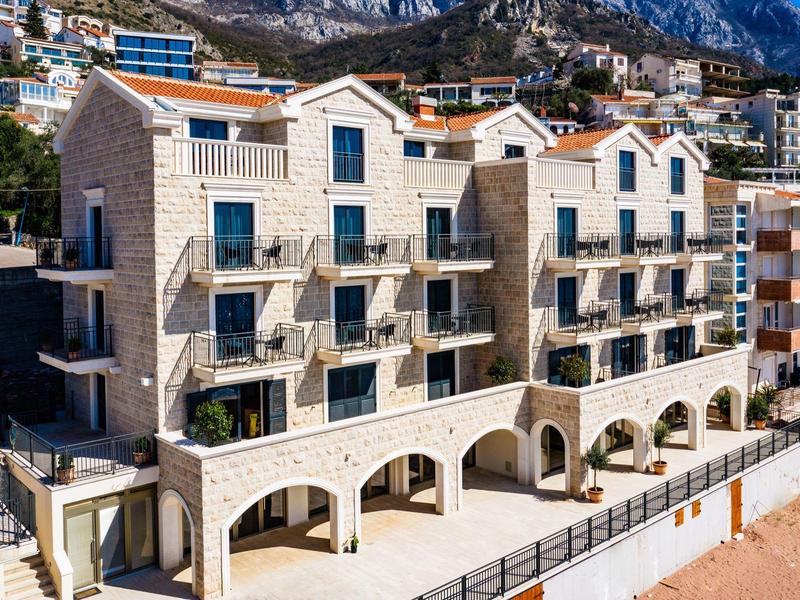 Modernes Steinhaus mit vielen Balkonen vor Bergkulisse und blauem Himmel.