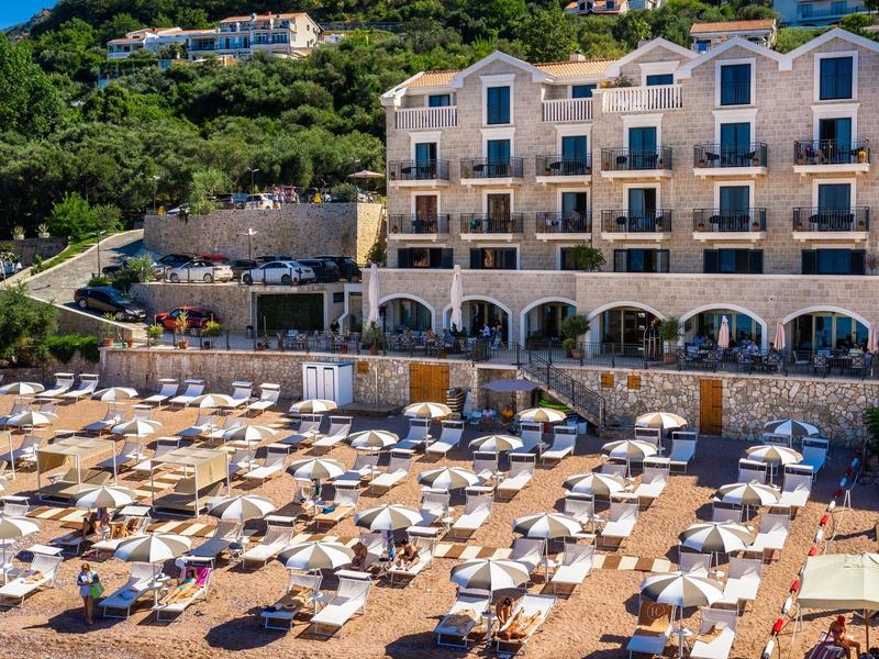 Strand mit vielen Liegen und Sonnenschirmen vor einem mehrstöckigen Hotel am grünen Hang.