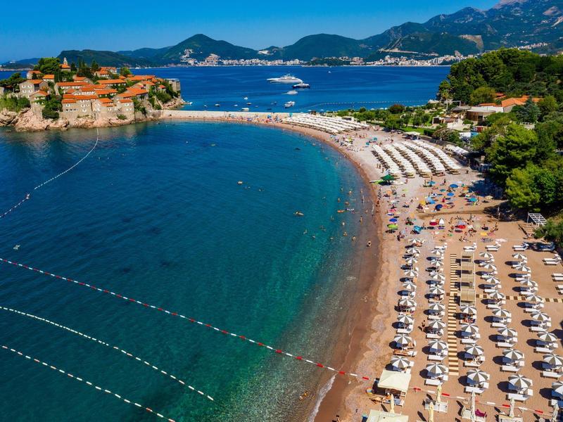 Sandstrand mit weißen Sonnenschirmen neben klarem Meer und kleiner Insel mit Gebäuden im Hintergrund.