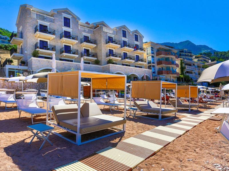 Strand mit Liegen und Pavillons vor mehrstöckigem Hotelgebäude unter blauem Himmel.