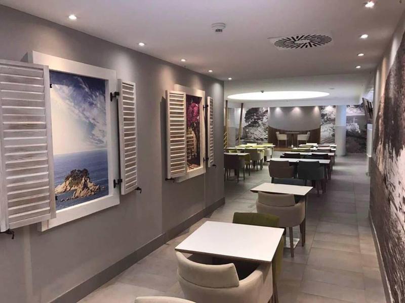 Moderner, heller Speisesaal mit rechteckigen Tischen, grünen Stühlen und dekorativen Bildern an grauen Wänden.