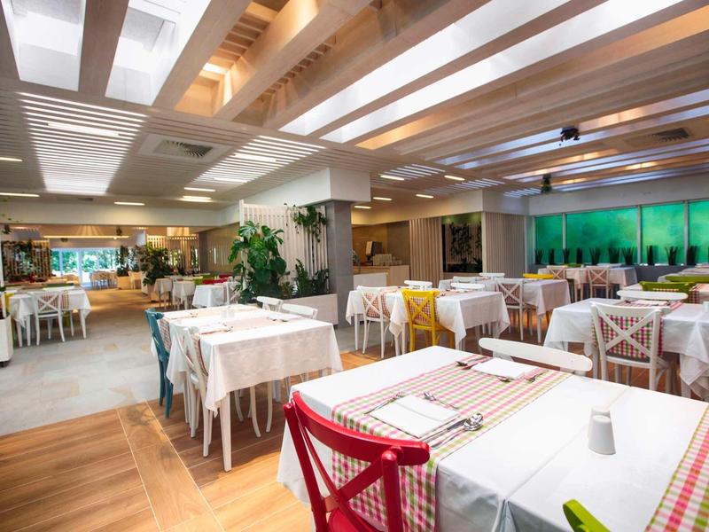 Helles Restaurant mit Holzdecken, weißen Tischdecken und roten Stühlen, Pflanzen und großer Fensterfront.