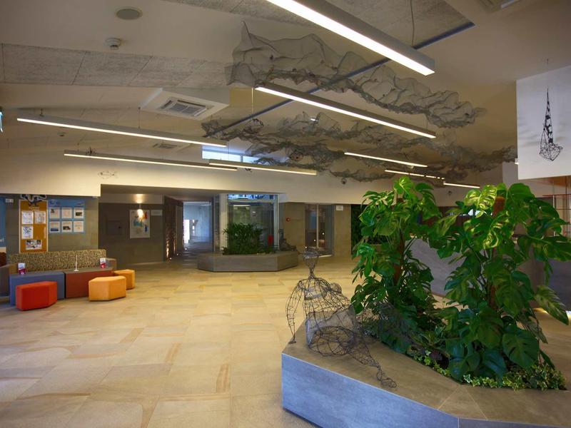 Moderner Flur mit hohen Decken, großen Pflanzen und bunten Sitzmöbeln in hellem Raum.