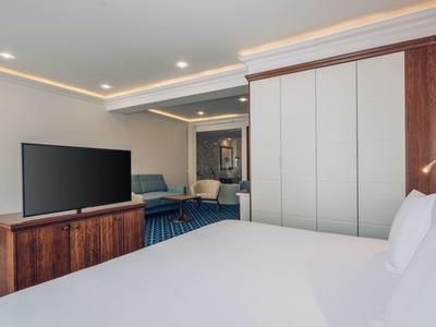 Helles Hotelzimmer mit großem Bett, TV auf einem Holzschrank und Sitzbereich im Hintergrund.